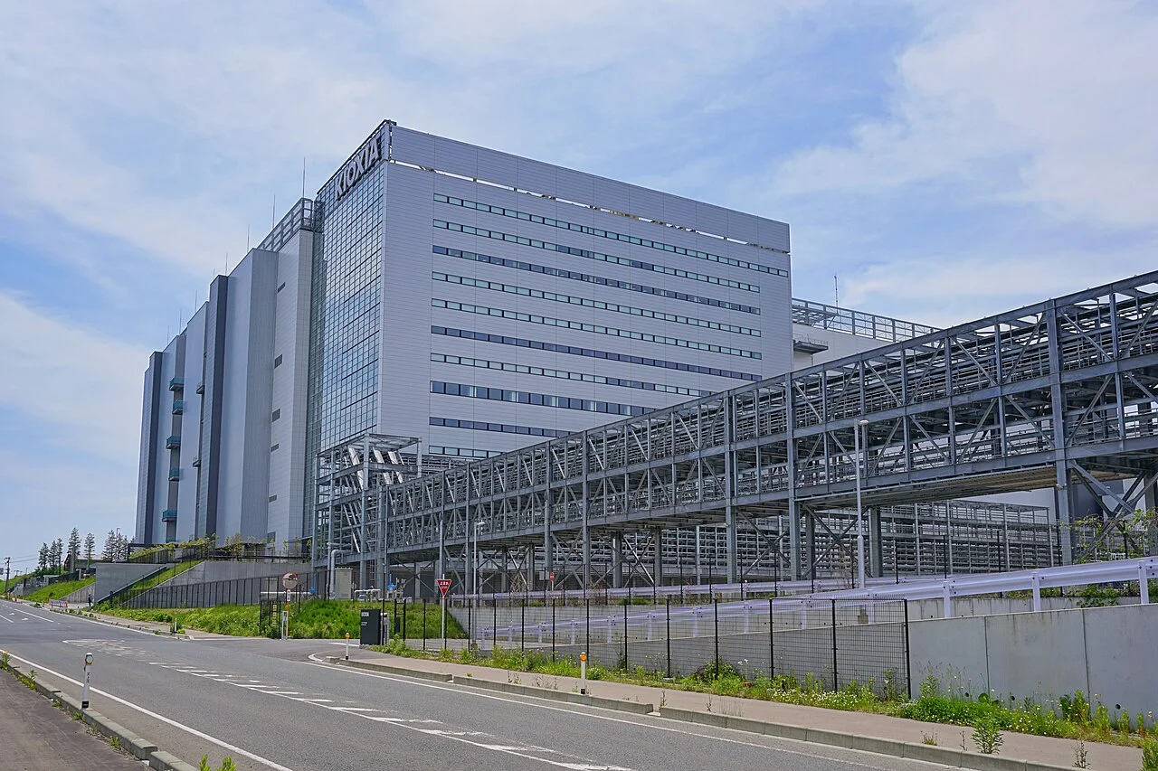 KIOXIAの文字が入った背の高い灰色の工場の建物と、手前に長く延びる鉄骨の渡り廊下。手前は道路で、空は青い