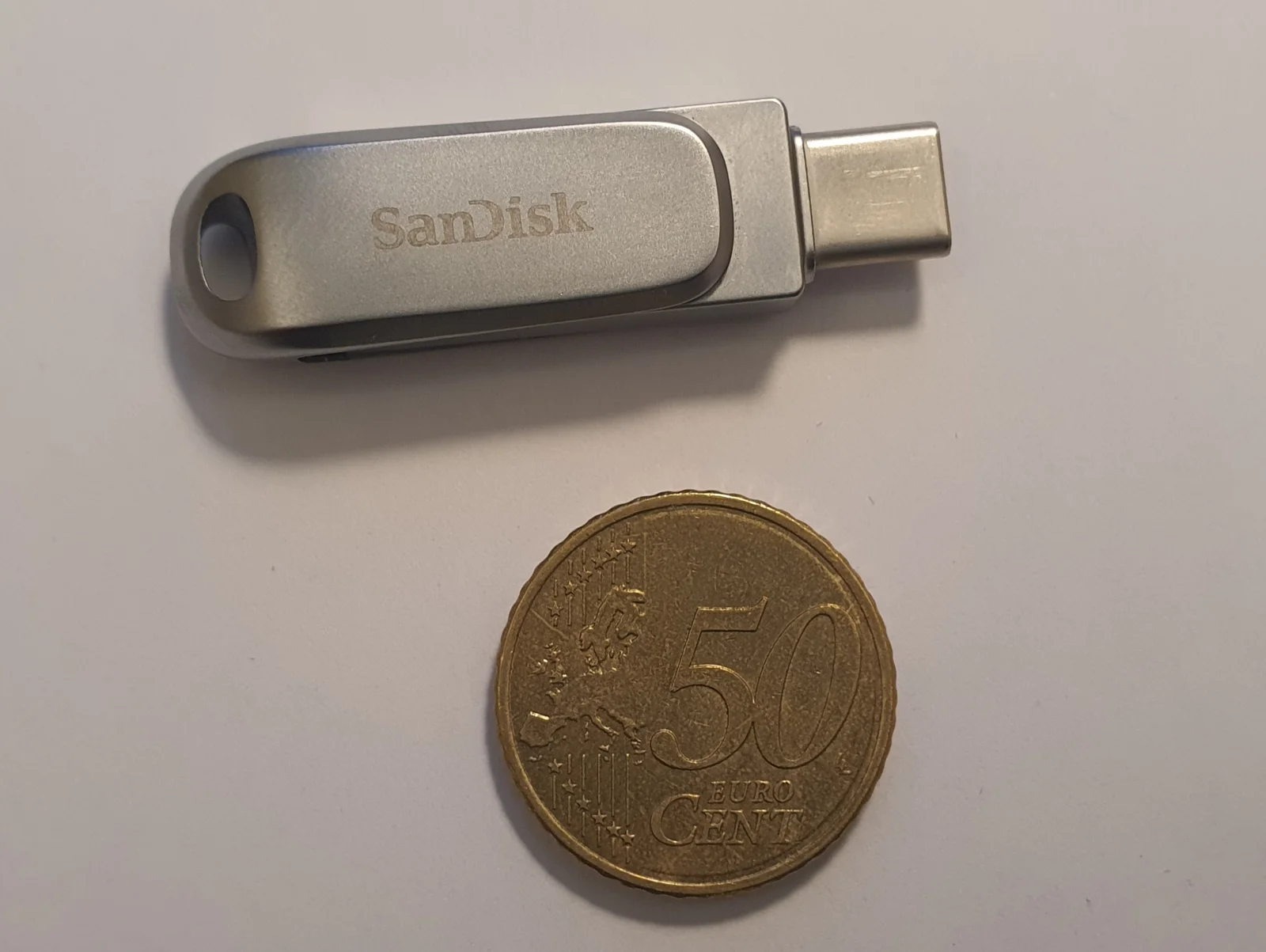 白い台の上に置いた銀色のUSBメモリ(SanDiskの刻印、端子はUSB Type-C)と、その下に並べた50ユーロセント硬貨。USBメモリの長さは硬貨の直径の2倍ほどしかない